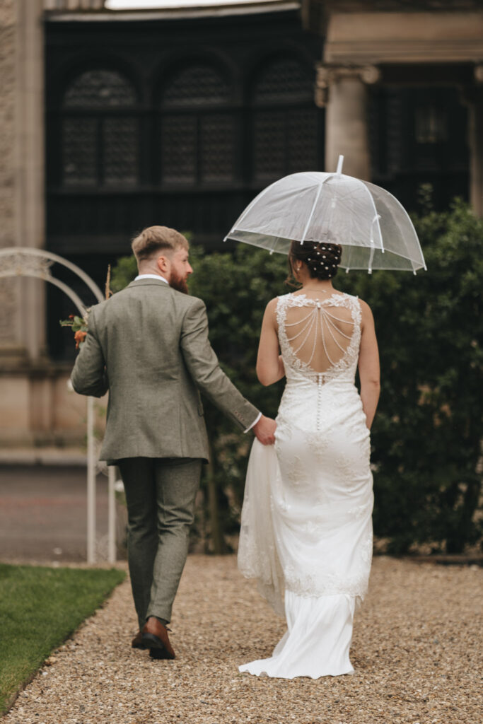A newlywed couple strolls down a gravel path at Bourton Hall. The groom, in a gray suit, holds the bride's hand as her white lace dress trails. She carries a transparent umbrella and wears a gown with an intricate lace back, perfect for their wedding photos by the historic building with arched windows and greenery. © Aimee Lince Photography