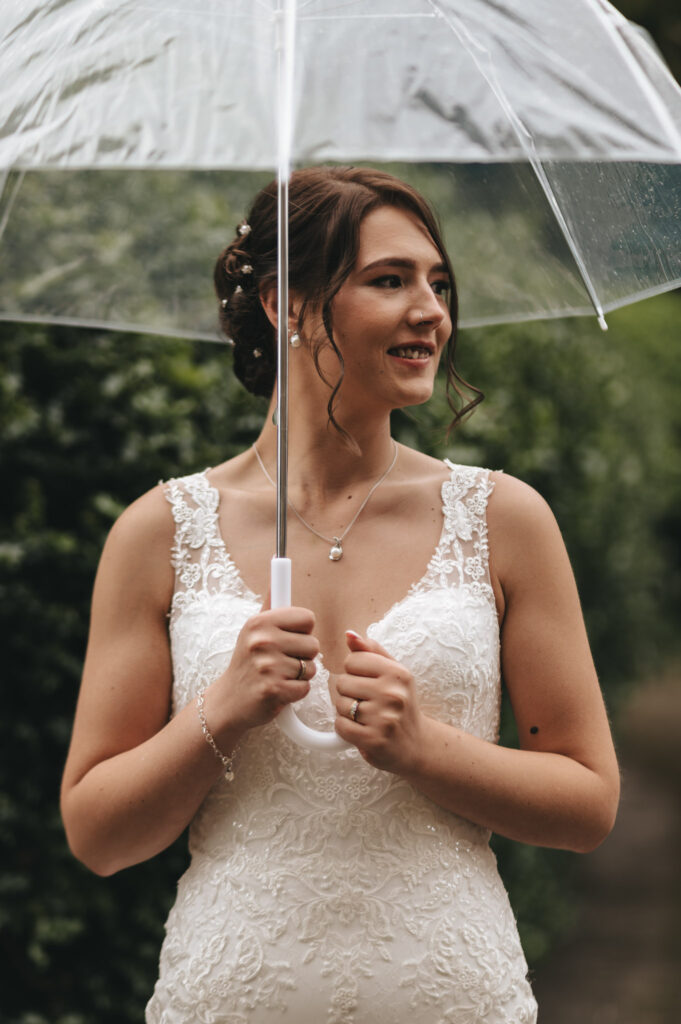 A woman in a white lace wedding dress poses under a clear umbrella at Bourton Hall. Her brown hair, styled in an updo with delicate flowers, complements her necklace and bracelet. The lush, green garden setting is beautifully captured by the wedding photographer, while she smiles despite the rain. © Aimee Lince Photography