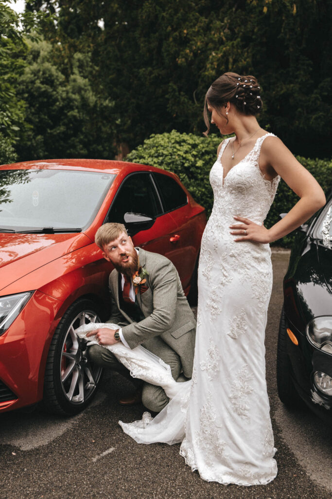 At a Bourton Hall Wedding, a groom in a gray suit playfully pretends to polish a tire on a red car while the bride, in her white lace dress, stands beside him with hands on her hips and smiles. Surrounded by greenery and a glimpse of another black car, the scene is captured beautifully by their wedding photographer. © Aimee Lince Photography
