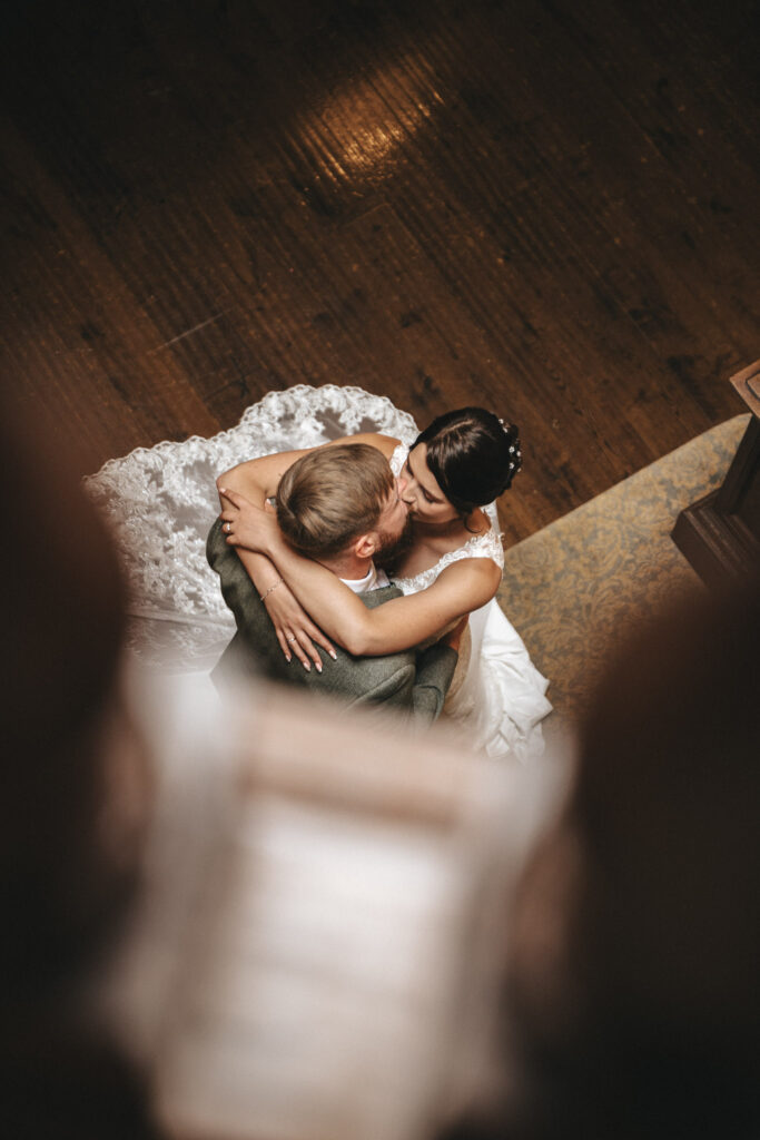 A bride and groom embrace closely while dancing on a wooden floor at Bourton Hall Wedding. The bride wears a white gown with lace details, and the groom is in a dark suit. Captured by a talented wedding photographer, the photo reveals an intimate moment with soft lighting and a blurred foreground. © Aimee Lince Photography