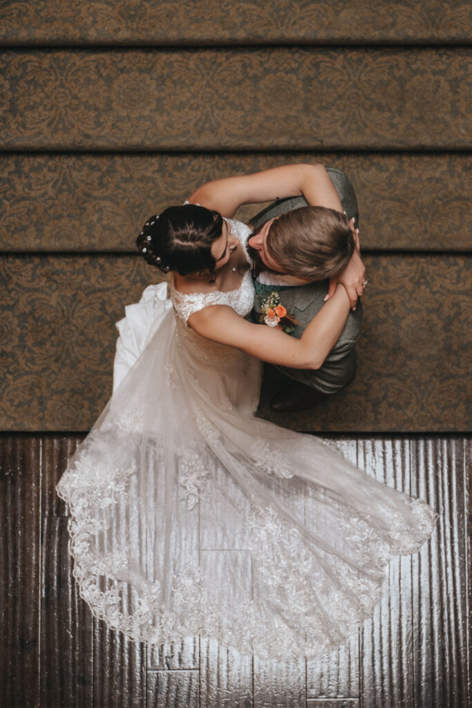 Overhead view of a bride in a lace wedding gown and a groom in a green suit embracing on the wooden floor, capturing the essence of Bourton Hall weddings. The bride's dress elegantly spreads out in a circular pattern near carpeted stairs, perfect for exquisite wedding photos. © Aimee Lince Photography