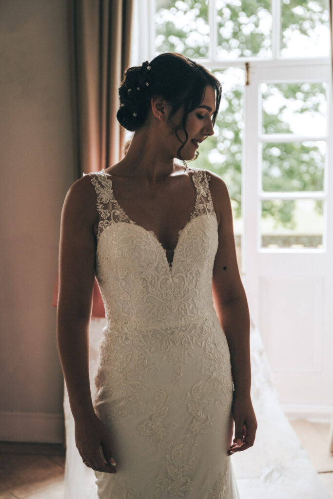 A bride stands indoors in a lace wedding dress with thin straps and a V-neckline, featuring intricate lace patterns. Her hair is styled in an elegant updo. Soft light filters in from a window with white frames at Bourton Hall, highlighting her serene expression. A blurred green landscape is visible outside—perfect for wedding photos. © Aimee Lince Photography