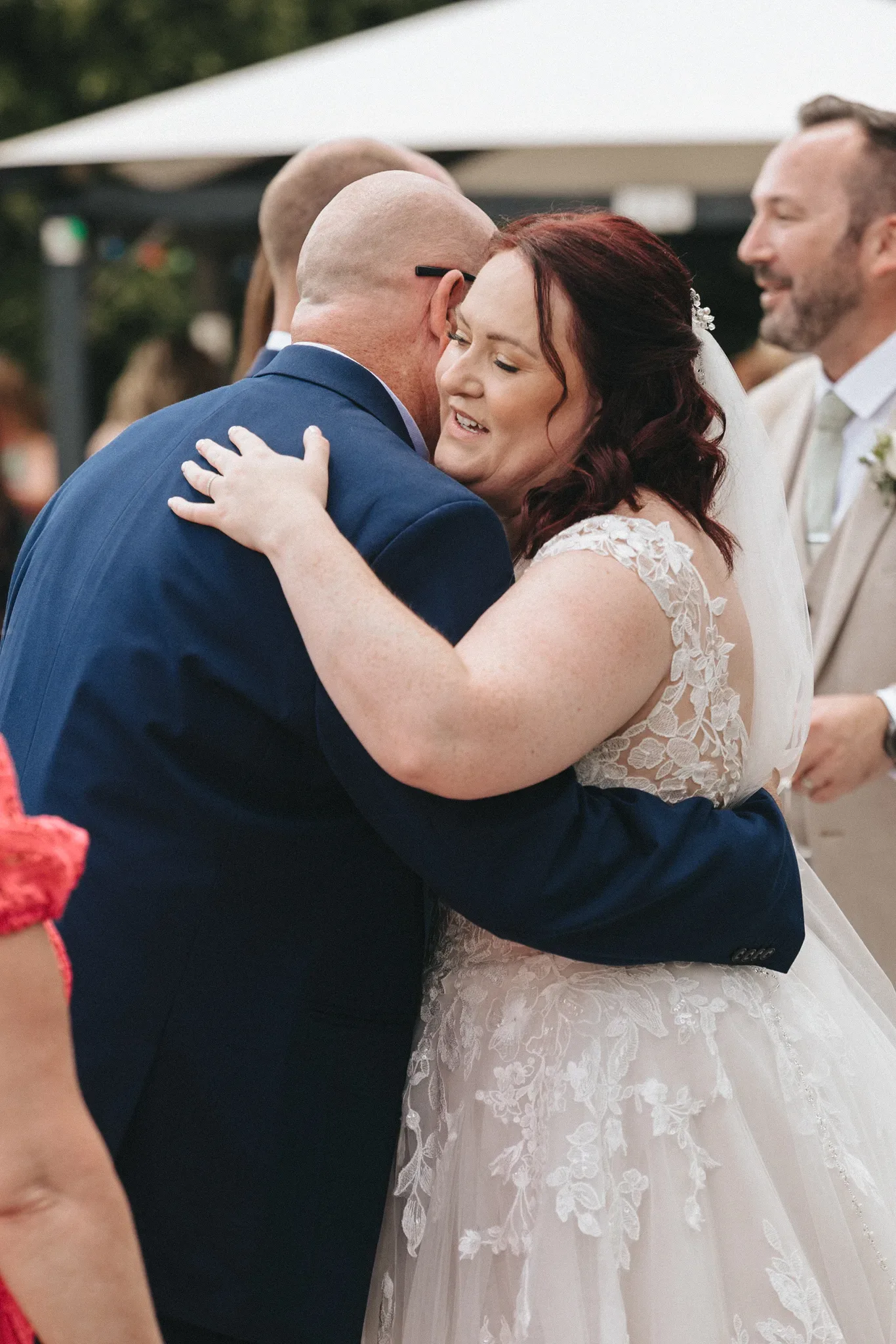 Bride in a white lace wedding dress with a veil, her red hair down, hugs a bald, bearded man wearing a blue suit. They are outdoors under a canopy, with blurred people in the background. The scene appears joyful and celebratory, embodying the essence of Hugs & High Fives on their special day. © Aimee Lince Photography