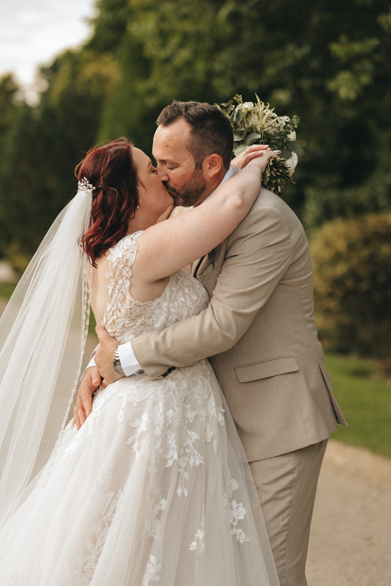 A couple kisses passionately outdoors, with lush greenery in the background. The bride, in a white lace wedding gown with a long veil, holds a bouquet. The groom is dressed in a beige suit. Wrapped in an embrace during their wedding at Brackenborough Hotel, the mood is romantic and serene. © Aimee Lince Photography