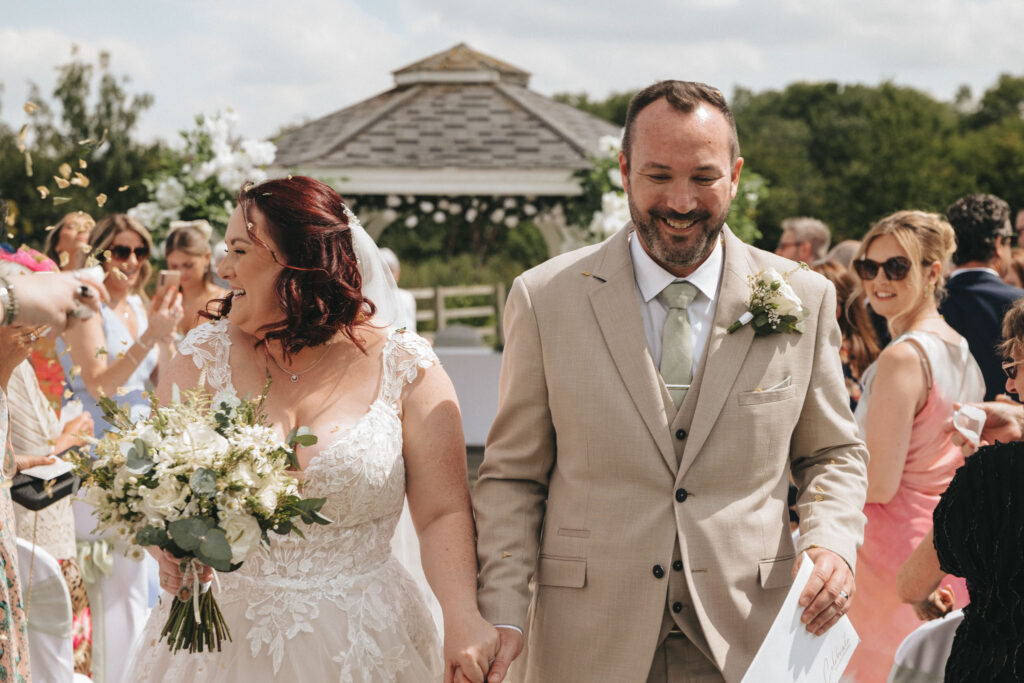 A bride and groom smile while walking down the aisle at their wedding at Brackenborough Hotel, as guests cheer and throw flower petals. The groom wears a beige suit, and the bride dons a white lace gown, holding a bouquet. A gazebo graces the background amidst lush greenery under a partly cloudy sky. © Aimee Lince Photography