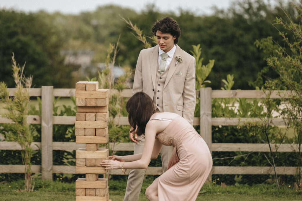 A woman in a pink dress carefully pulls a block from a large wooden Jenga tower on the grass during the wedding at Brackenborough Hotel. A man in a beige suit watches her, surrounded by greenery and a wooden fence under the cloudy sky. © Aimee Lince Photography