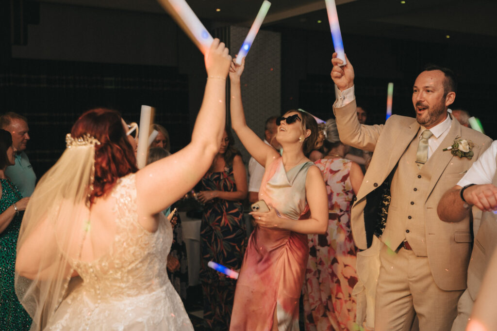 At a lively wedding at Brackenborough Hotel, a bride in a lace gown twirls her colorful glow stick. Nearby, a woman donning sunglasses and pink attire joins in the fun, glow stick in hand. A man in a beige suit dances alongside them as other party-goers create an unforgettable scene. © Aimee Lince Photography