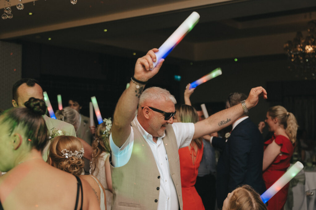 At a lively wedding at Brackenborough Hotel, the party scene buzzes as people dance energetically. A man in a beige vest and sunglasses raises glowing sticks, surrounded by others in formal attire. Bright lights and a chandelier enhance the festive atmosphere, with tattoos visible on his arm. © Aimee Lince Photography