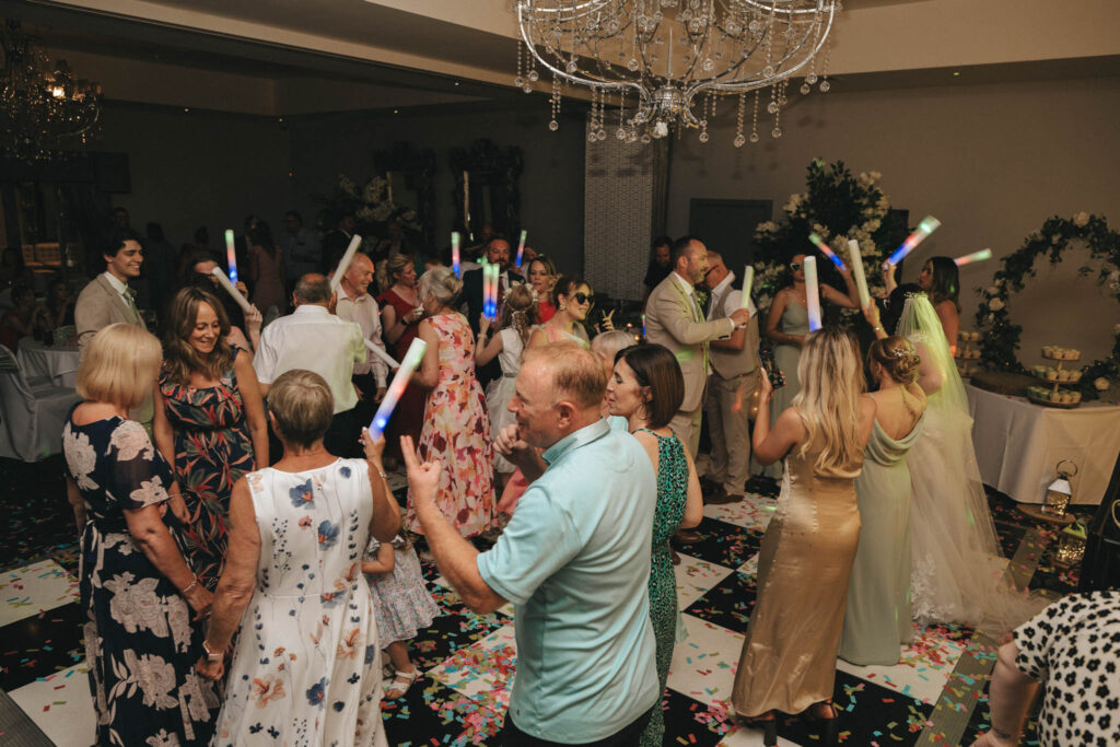 A vibrant wedding reception at Brackenborough Hotel has guests dancing and holding glow sticks. The dance floor is covered in confetti, with people joyfully interacting under a large, ornate chandelier. The bride, in a flowing white dress, mingles among guests, creating a lively and celebratory atmosphere. © Aimee Lince Photography