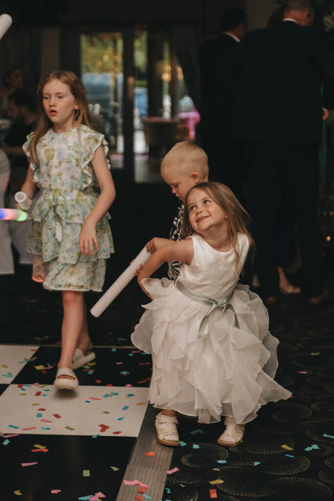 Three children in a festive setting at a wedding at Brackenborough Hotel. A girl in a floral dress walks holding a white tube, a blond boy focuses downward, and a smiling girl in white twirls joyfully. Colorful confetti covers the black and white floor, suggesting a celebratory atmosphere. © Aimee Lince Photography