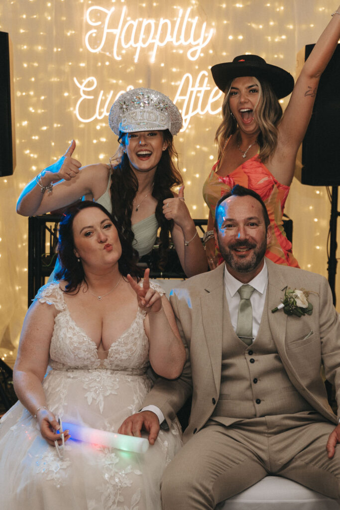 A joyful wedding celebration at Brackenborough Hotel, with two seated people: the bride in a white lace dress and the groom in a tan suit. Behind them, two standing guests wear playful hats—one silver and one black. A neon sign reads "Happily Ever After." Everyone is smiling and making playful gestures. © Aimee Lince Photography