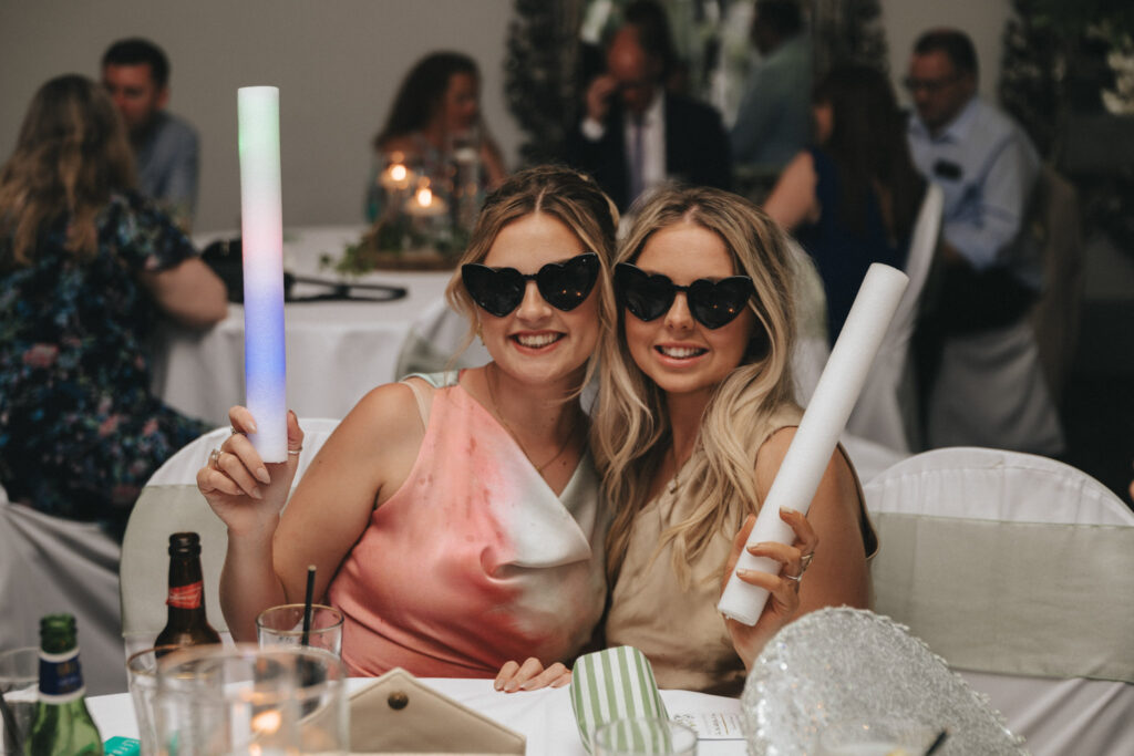 Two women wearing heart-shaped sunglasses smile at the camera, seated at a decorated table during a wedding at Brackenborough Hotel. Both hold glow sticks—one multicolored, the other white. Drinks and a shiny handbag are on the table as other guests mingle in the softly lit room. © Aimee Lince Photography