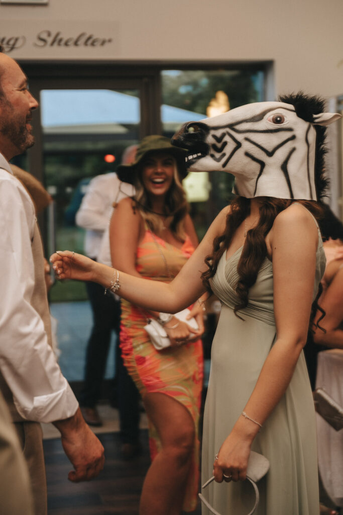 At a wedding at Brackenborough Hotel, a woman in a light green dress and zebra mask chats with a man in a white shirt and beige vest. In the background, another woman in a colorful dress and hat beams with joy. Sunlight spills through large windows showcasing lush greenery outside. © Aimee Lince Photography