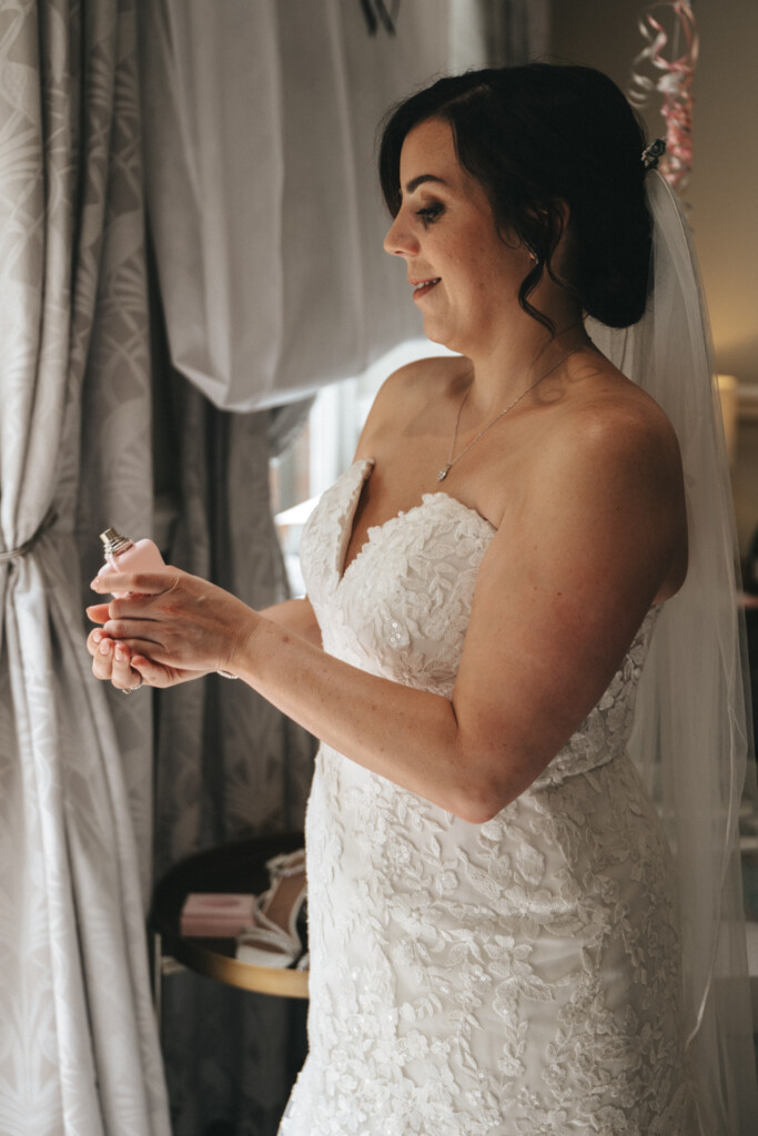 A bride in a white lace strapless wedding dress holds a small pink perfume bottle near a window. She has dark hair styled in loose curls and wears a necklace and veil. Behind her, a table with shoes and decorations is visible. The room has patterned curtains and soft lighting. © Aimee Lince Photography