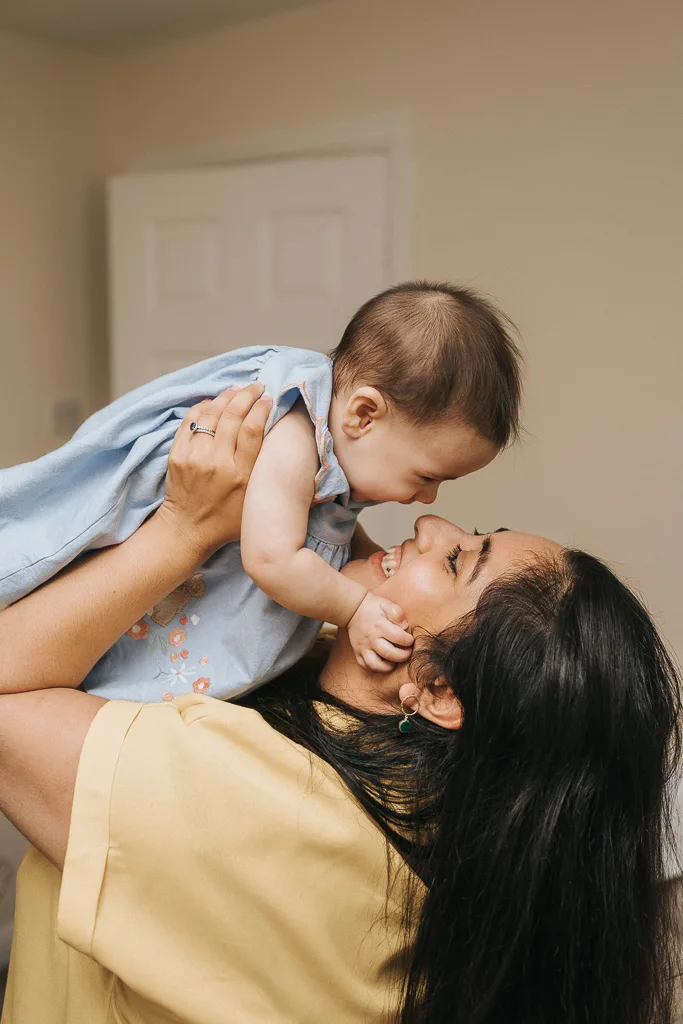 A woman with long dark hair smiles warmly as she lifts a baby in a blue, embroidered dress above her head. The baby, with short hair, gazes down joyfully at her. Set against a beige indoor background with a white door, the scene captures their shared delight and tender bond. © Aimee Lince Photography - Wedding photographer in Lincolnshire, Yorkshire & Nottinghamshire