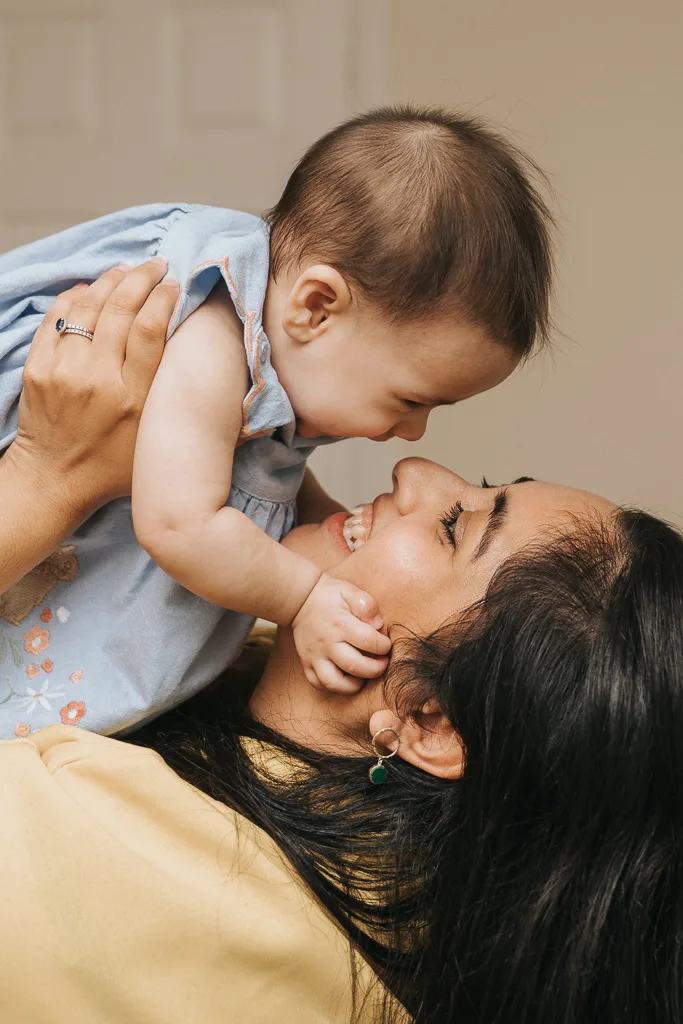 A woman with long black hair lies on a bed, smiling and lifting a baby in a light blue dress. The baby leans forward, nosing her gently, their smiles capturing the pure joy of the moment. The softly blurred background highlights their delightful interaction. © Aimee Lince Photography - Wedding photographer in Lincolnshire, Yorkshire & Nottinghamshire