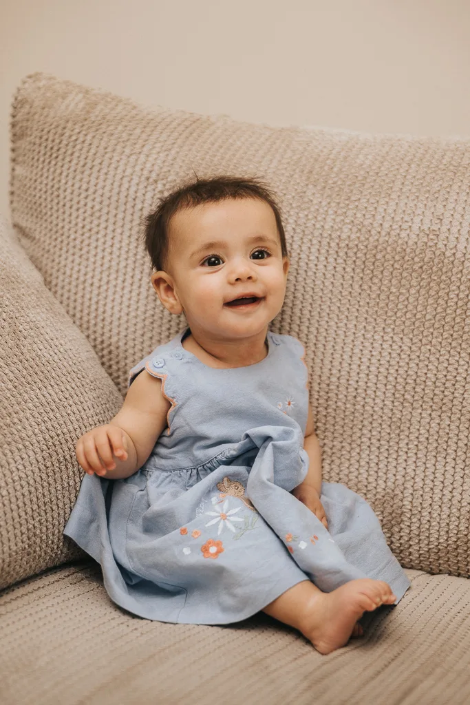 A baby with short dark hair sits on a textured beige couch, wearing a light blue dress embroidered with orange and white flowers. The baby is looking up with a slight smile, holding the dress in one hand, exuding a curious and cheerful expression. © Aimee Lince Photography - Wedding photographer in Lincolnshire, Yorkshire & Nottinghamshire