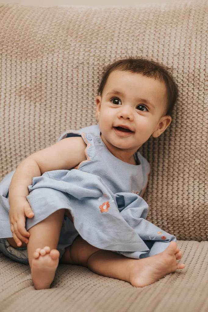 A smiling baby sits on a textured beige couch, wearing a light blue dress with orange floral embroidery. The baby has short dark hair and bare feet, with one hand resting on the couch. The scene is warmly lit, creating a cozy and cheerful atmosphere perfect for this adorable little one. © Aimee Lince Photography - Wedding photographer in Lincolnshire, Yorkshire & Nottinghamshire