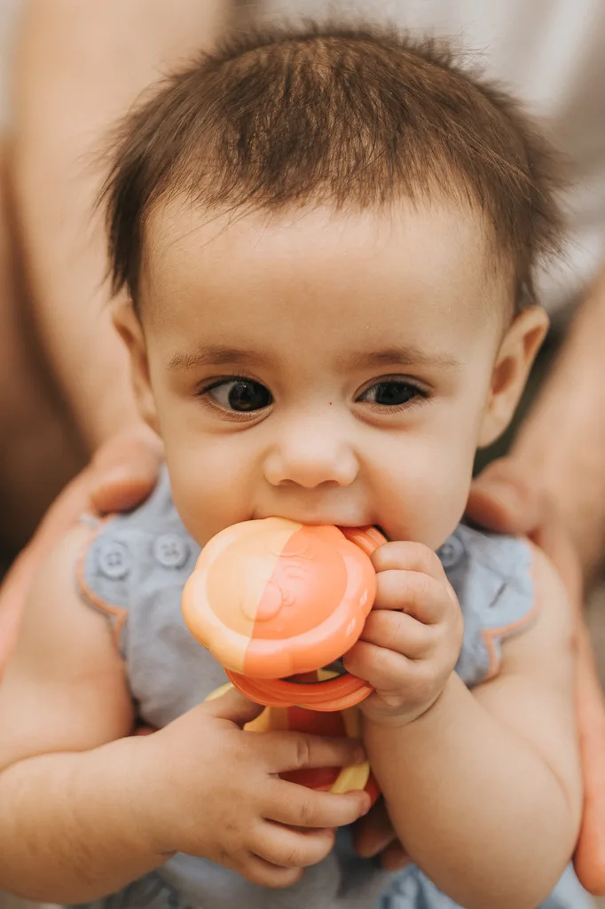 A baby with short, dark hair wearing a light blue sleeveless outfit is adorably chewing on a round orange and yellow teething toy. The infant sits comfortably with an adult's hands gently supporting them from behind, all set against a softly blurred background. © Aimee Lince Photography - Wedding photographer in Lincolnshire, Yorkshire & Nottinghamshire