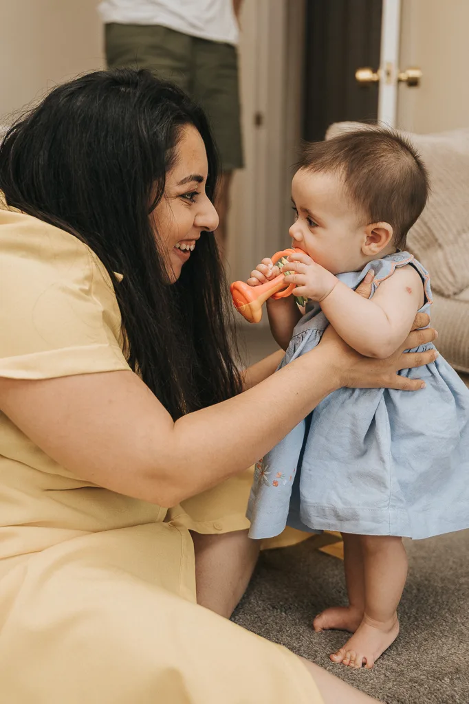 A woman in a yellow dress kneels on the carpeted floor, smiling at a baby in a light blue dress who is standing and holding an orange rattle. The scene is indoors, with open doors in the background and another person partially visible nearby. © Aimee Lince Photography - Wedding photographer in Lincolnshire, Yorkshire & Nottinghamshire