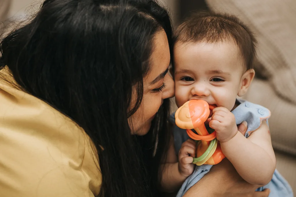 A smiling baby in a blue outfit holds a colorful orange and green toy, while a woman with long dark hair leans in affectionately, wearing a yellow top. They are indoors, with a neutral background, creating an undeniably heartwarming and joyful baby scene. © Aimee Lince Photography - Wedding photographer in Lincolnshire, Yorkshire & Nottinghamshire