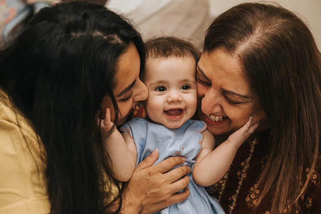 A baby in a blue dress smiles while being embraced by two women. One kisses its cheek as the other snuggles close, both smiling joyfully. The heartwarming scene radiates warmth and affection. © Aimee Lince Photography - Wedding photographer in Lincolnshire, Yorkshire & Nottinghamshire