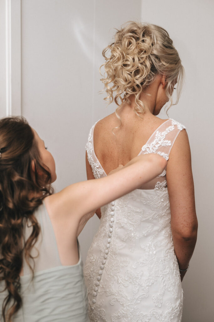 A bride in a lace wedding dress with a buttoned back is being assisted by a young girl. Her hair is styled in curls as she stands with her back to the viewer. Captured by a skilled Louth wedding photographer, the scene unfolds with the young girl, in pale green, carefully fastening the buttons. © Aimee Lince Photography - Wedding photographer in Lincolnshire, Yorkshire & Nottinghamshire