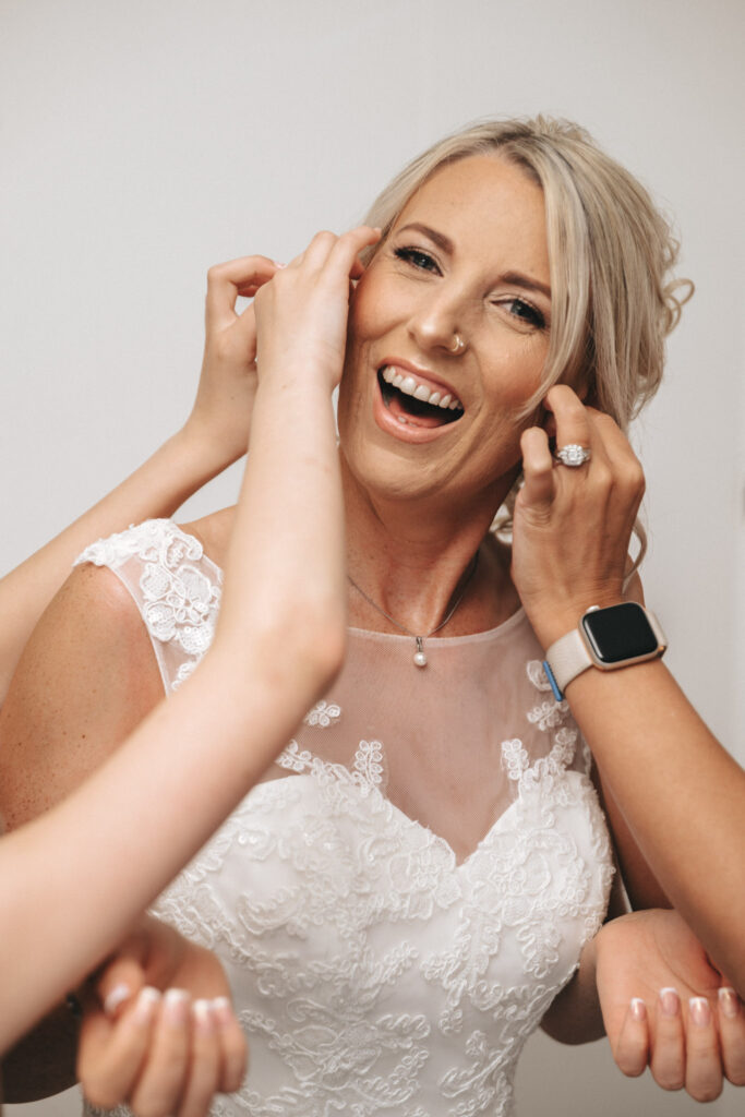 A smiling bride with blonde hair is adorned in a white lace wedding dress. As she prepares for her Louth wedding, two people adjust her hair and earrings. She sports a nose ring, necklace, and a ring on her left hand. One helper wears a smartwatch, while the bride radiates joy with her wide smile. © Aimee Lince Photography