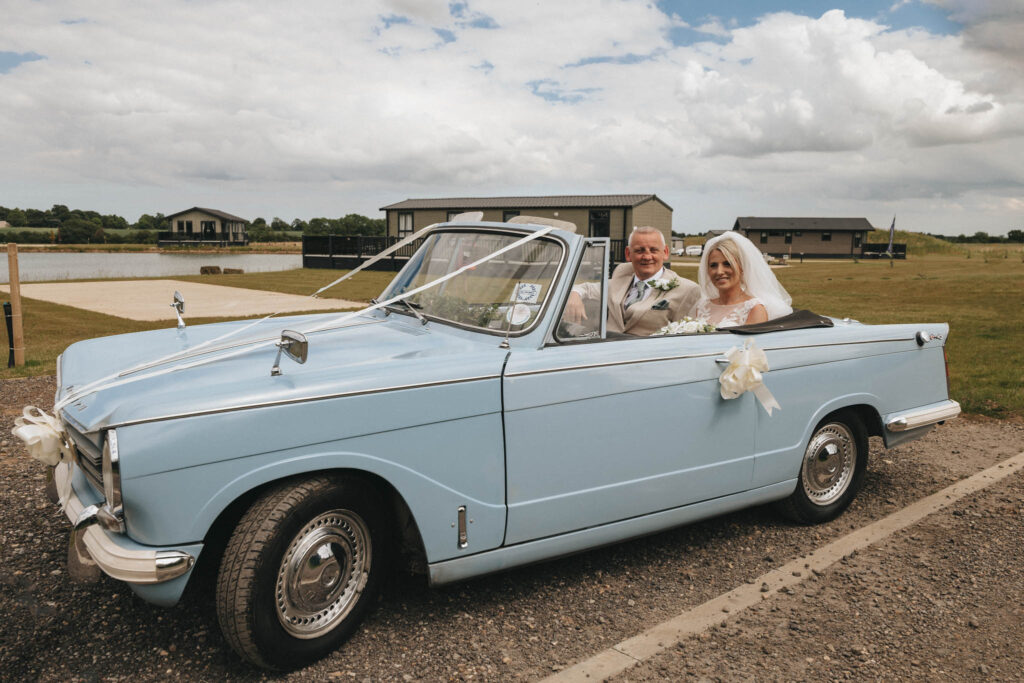 An elderly couple sits in a light blue vintage convertible car adorned with white ribbons, captured on a cloudy Louth day. The woman in a white dress and veil beams beside the man in a grey suit. Parked on a gravel road near a grassy field, they celebrate their perfect wedding moment. © Aimee Lince Photography