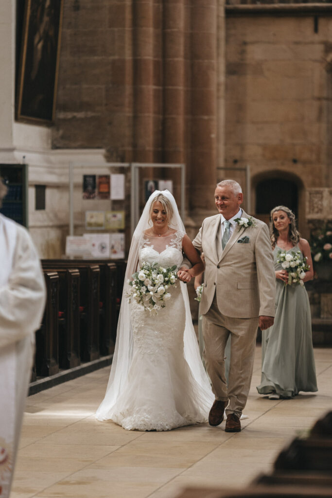 A bride in a white dress and veil holds a bouquet of white flowers, walking down the aisle of a Louth church with an older man in a beige suit. A bridesmaid in a light green dress follows, holding a similar bouquet. The church interior is adorned with stone walls and wooden pews. © Aimee Lince Photography