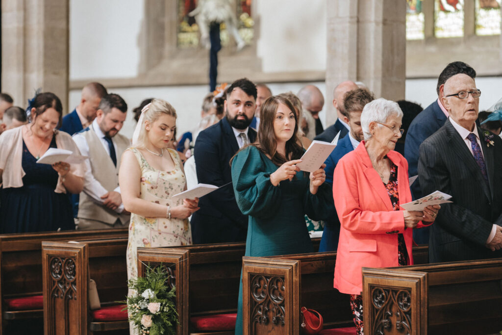 A congregation stands in a church in Louth, holding programs for a wedding. The scene includes people of various ages dressed formally. Wooden pews are visible, and stained glass windows cast colorful light over bouquets of flowers decorating the pews. © Aimee Lince Photography