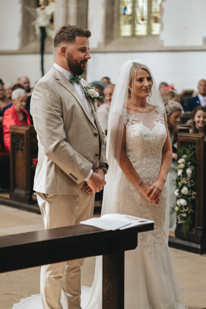 A bride and groom stand at the altar during a beautiful Louth wedding ceremony. The bride wears a lace wedding dress with a veil, and the groom is in a beige suit with a bow tie. They are surrounded by seated guests in a church, with stained glass windows and green floral arrangements adorning the backdrop. © Aimee Lince Photography