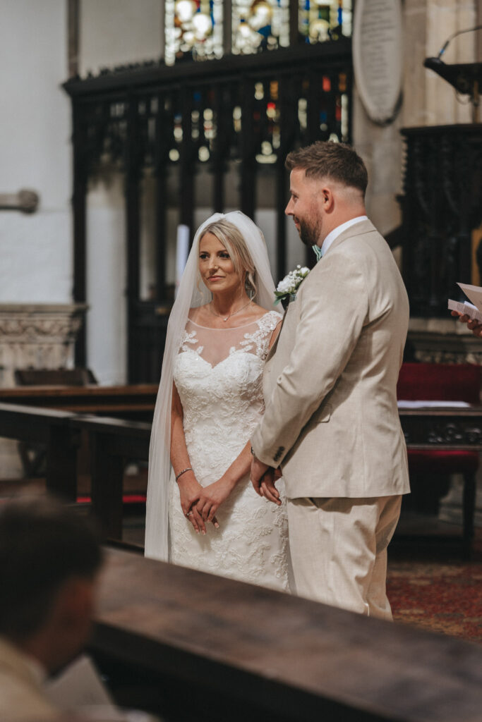 A bride in a white lace gown and veil stands beside a groom in a beige suit during their Louth wedding, facing the officiant. The church, adorned with stained glass and dark wood accents, frames the solemn couple as they look forward attentively. © Aimee Lince Photography