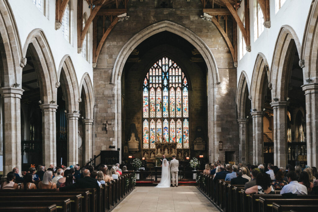 A couple stands at the altar of a grand church during their Louth wedding ceremony. The congregation is seated in wooden pews along the central aisle. Above, a large, colorful stained glass window is illuminated, while high arches and stone architecture complete the breathtaking scene. © Aimee Lince Photography