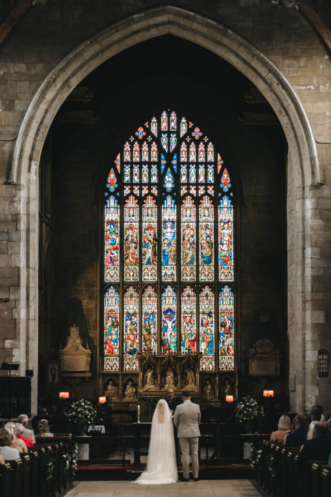 A bride and groom stand at the altar in a historic church in Louth, with a large, colorful stained glass window. The arched ceiling and stone walls frame the ceremony of this beautiful Louth wedding. Guests sit in pews on both sides, facing the couple as natural light filters through the glass. © Aimee Lince Photography