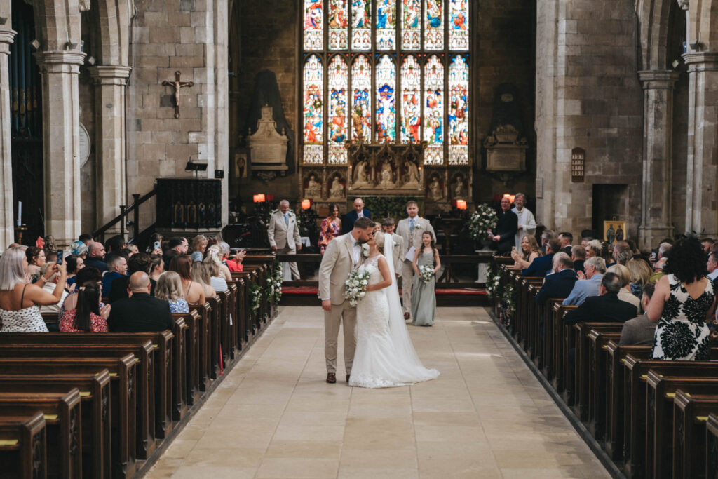 At a charming Louth wedding, the bride and groom share a kiss at the altar in a large stone church. Guests fill the pews, some taking photos. Large stained glass windows and candles adorn the background. The aisle is lined with flowers, creating a brightly lit romantic scene. © Aimee Lince Photography