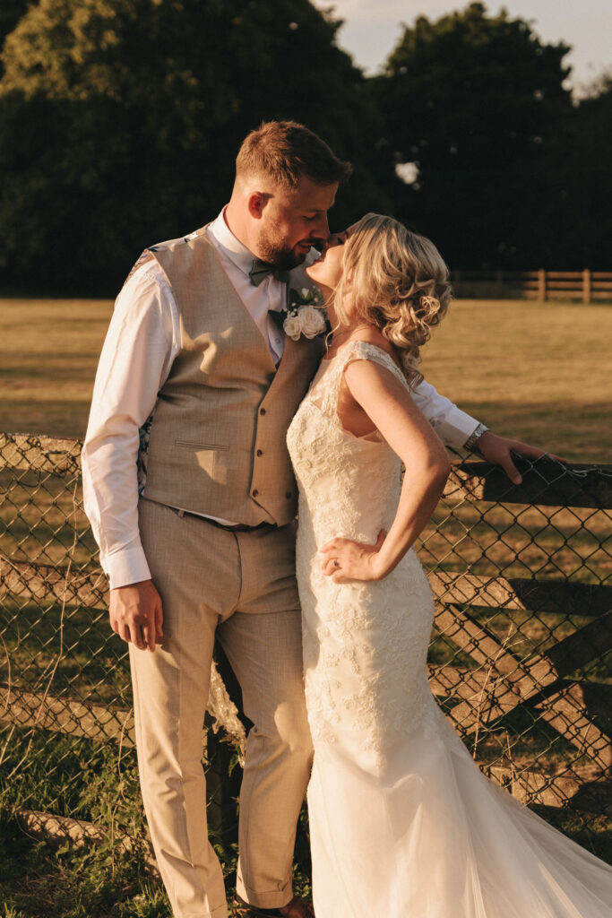 A bride and groom share an intimate moment outdoors during their Louth wedding. The groom, in a beige vest and white shirt, leans against a rustic fence as the bride, in a lace dress, gently leans towards him. They are surrounded by a sunlit field with trees in the background, creating a romantic atmosphere. © Aimee Lince Photography