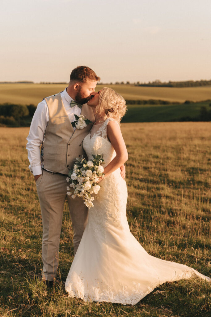 In a sunlit field in Louth, a bride and groom share a kiss. The groom dons a beige vest and trousers with a white shirt, while the bride dazzles in her lace dress, clutching a bouquet of white flowers. Rolling green hills under a clear sky frame this idyllic wedding scene. © Aimee Lince Photography