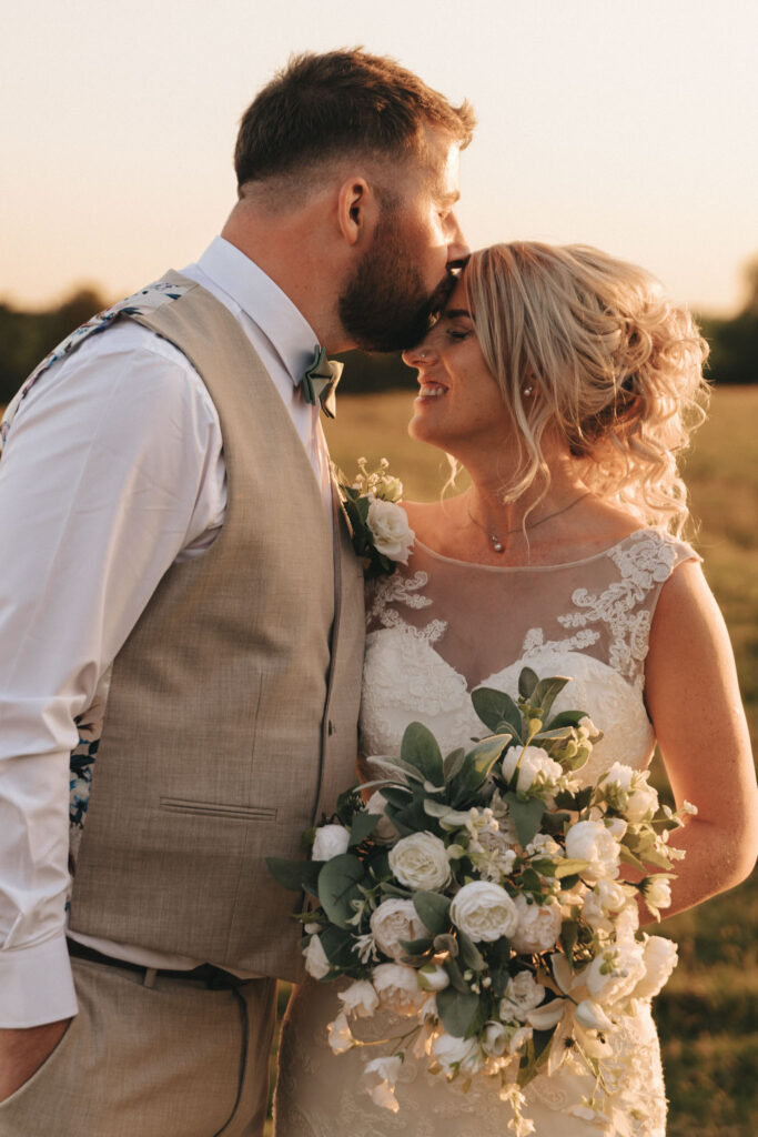 In the sunlit fields of Louth, a groom in a beige vest kisses the forehead of his smiling bride dressed in lace. Her bouquet of white flowers and greenery adds to their romantic wedding scene, enveloped in a soft, golden glow. © Aimee Lince Photography