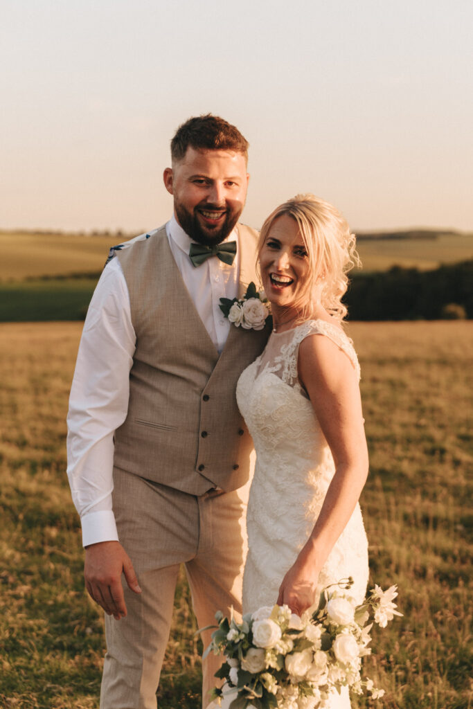 A smiling couple stands in a sunlit field at their Louth wedding. The man wears a beige vest and white shirt with a bow tie, while the woman dons a lace wedding dress and holds a bouquet of white flowers. Rolling hills under a clear sky cast a golden glow over the romantic scene. © Aimee Lince Photography