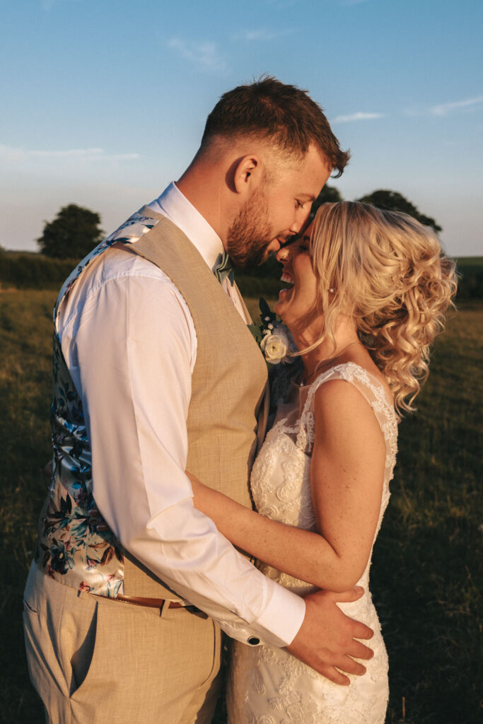 In a sunlit field, during their Louth wedding, a couple stands close in their elegant attire. The groom dons a tan suit with a floral vest, while the bride radiates in her lace dress. They smile as their foreheads touch, the sunset casting a warm glow with trees silhouetted behind them. © Aimee Lince Photography