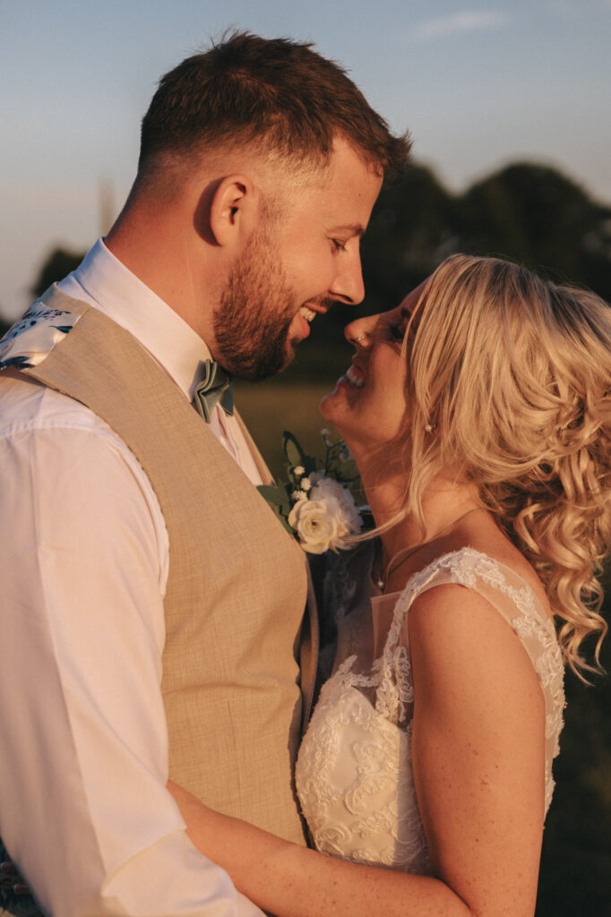 A bride and groom embrace outdoors at sunset during their Louth wedding. The groom wears a beige vest and white shirt, and the bride is in a lace wedding dress. They smile at each other under the warm glow of the evening sun, with blurred greenery and a clear sky in the background. © Aimee Lince Photography