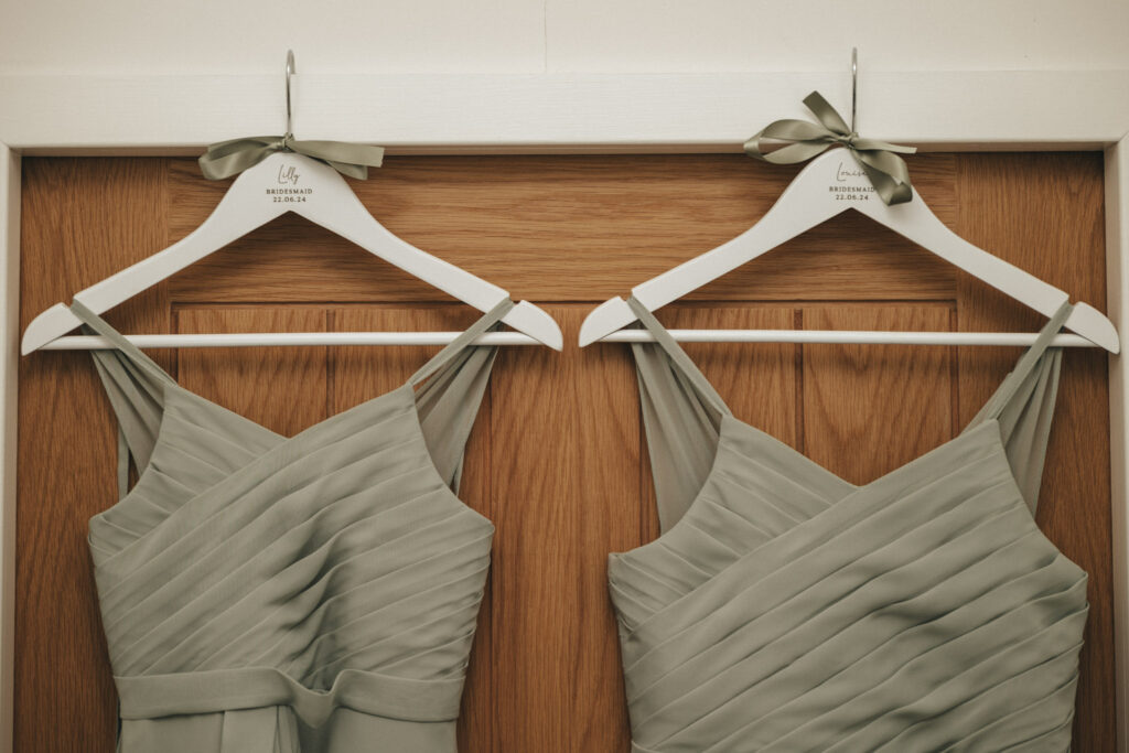Two sage green pleated dresses hang on white wooden hangers against a wooden door, perfect for a Louth wedding. Each hanger features a matching sage green ribbon tied into a bow at the hook. The pleats create a diagonal pattern, and the hangers display the logo "Lily" with smaller text below. © Aimee Lince Photography