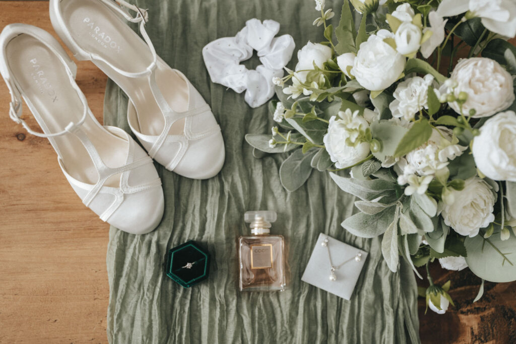 A flat lay of Louth wedding items includes white shoes, a white scrunchie, a bouquet of white and green flowers, a small green jewelry box with a ring, perfume, and a delicate necklace in an open box, all elegantly arranged on textured green cloth. © Aimee Lince Photography