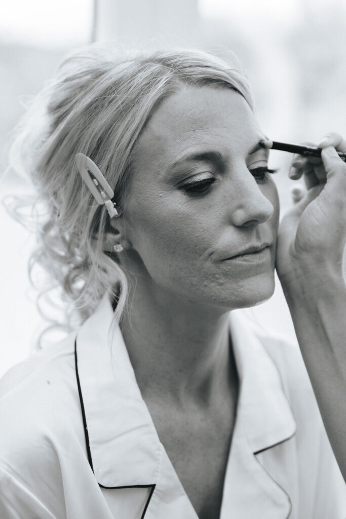 A black-and-white image captures a bride in Louth having her wedding makeup applied. Her light hair is pinned back with a clip, complementing the collared shirt with dark piping. With closed eyes, she exudes calm as the artist delicately applies eyeliner against the softly blurred background. © Aimee Lince Photography