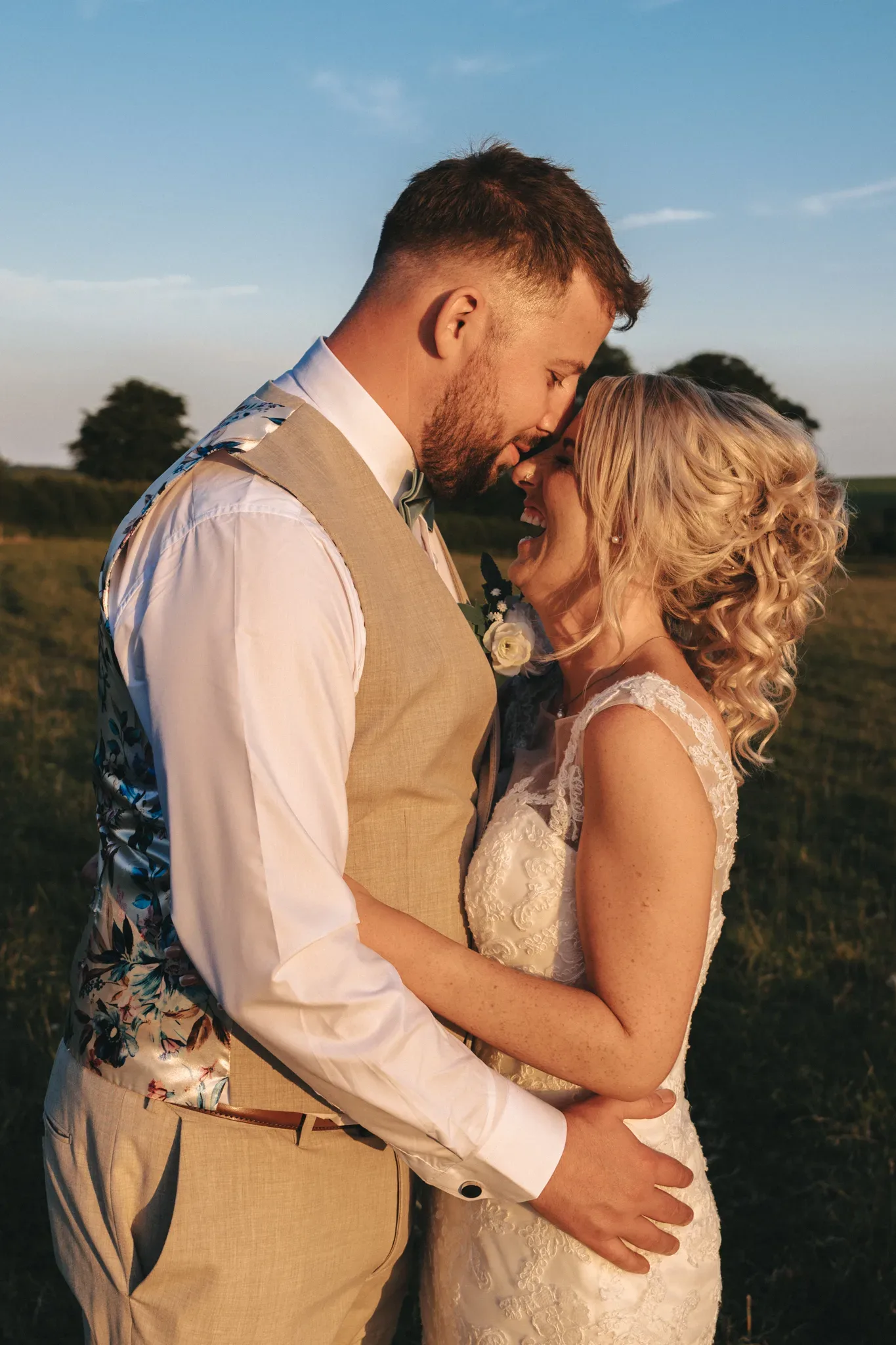 A newly married couple stands in a grassy field at sunset, embracing each other. The groom wears a beige vest with a floral pattern on the back, a white shirt, and a floral tie. The bride wears a white lace dress, her blonde hair styled in curls. They smile with their foreheads touching. © Aimee Lince Photography