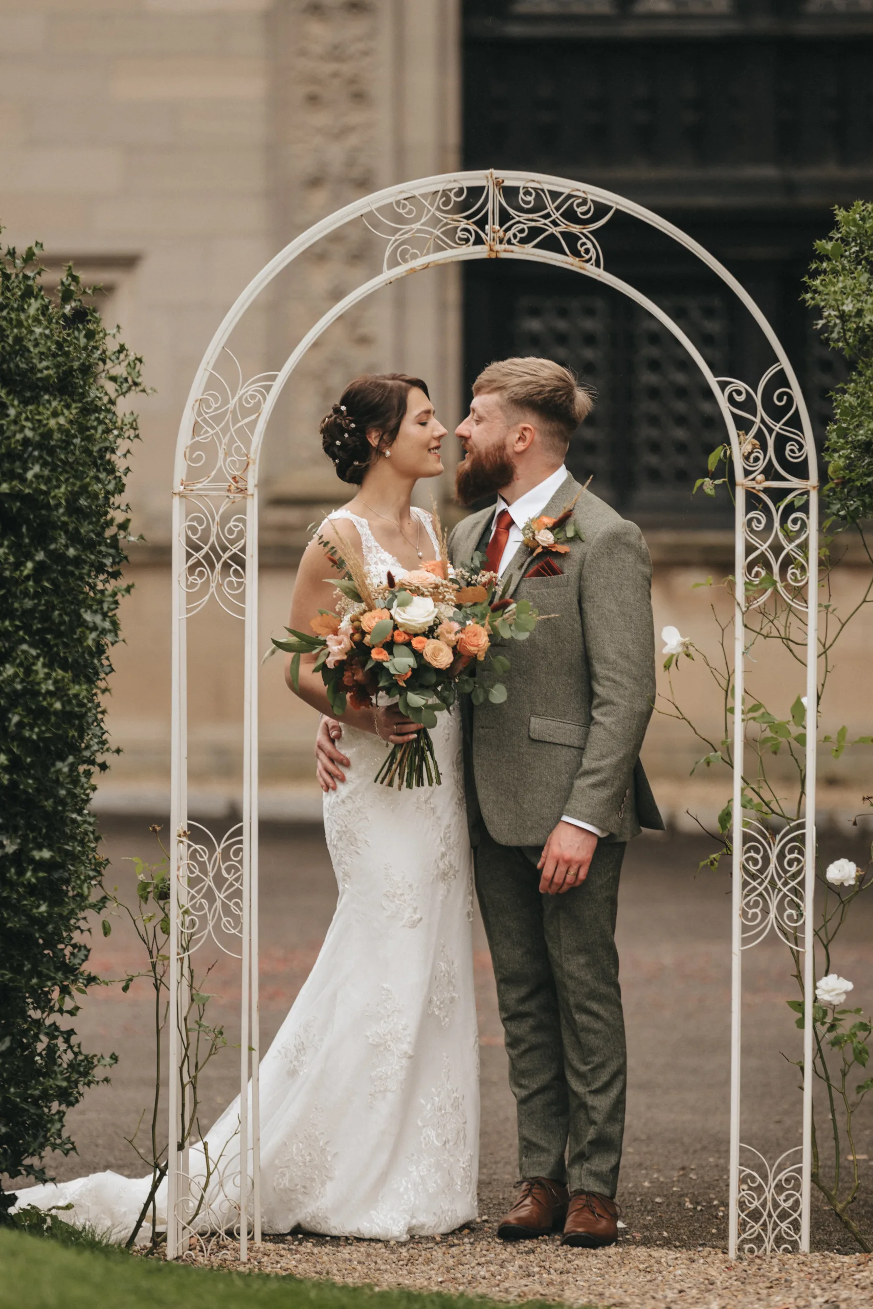 A couple stands under a white wrought iron arch, surrounded by green bushes. The bride wears a white lace gown and holds a bouquet of orange and white flowers. The groom wears a gray suit with a brown tie and boutonniere. They gaze into each other's eyes, smiling affectionately. © Aimee Lince Photography