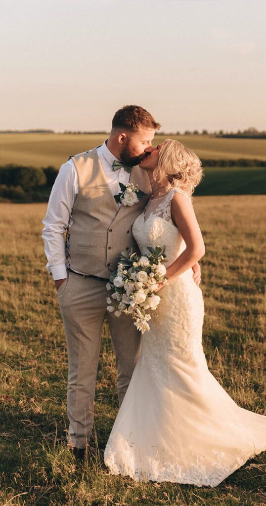 In the serene embrace of a Louth wedding, a couple stands in a grassy field at sunset. The groom, in a beige vest and pants with a white shirt, and the bride, in a lace gown holding white flowers, are about to kiss amid rolling hills and trees. © Aimee Lince Photography