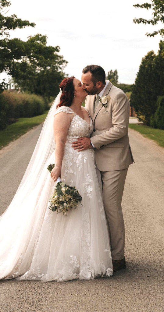 A couple shares a kiss on a tree-lined road during their wedding at Brackenborough Hotel. The bride, in a lace wedding dress with a long veil, holds a bouquet of white and green flowers. The groom dons a beige suit with a white shirt and boutonniere. Lush greenery adorns the backdrop beneath the partly cloudy sky. © Aimee Lince Photography