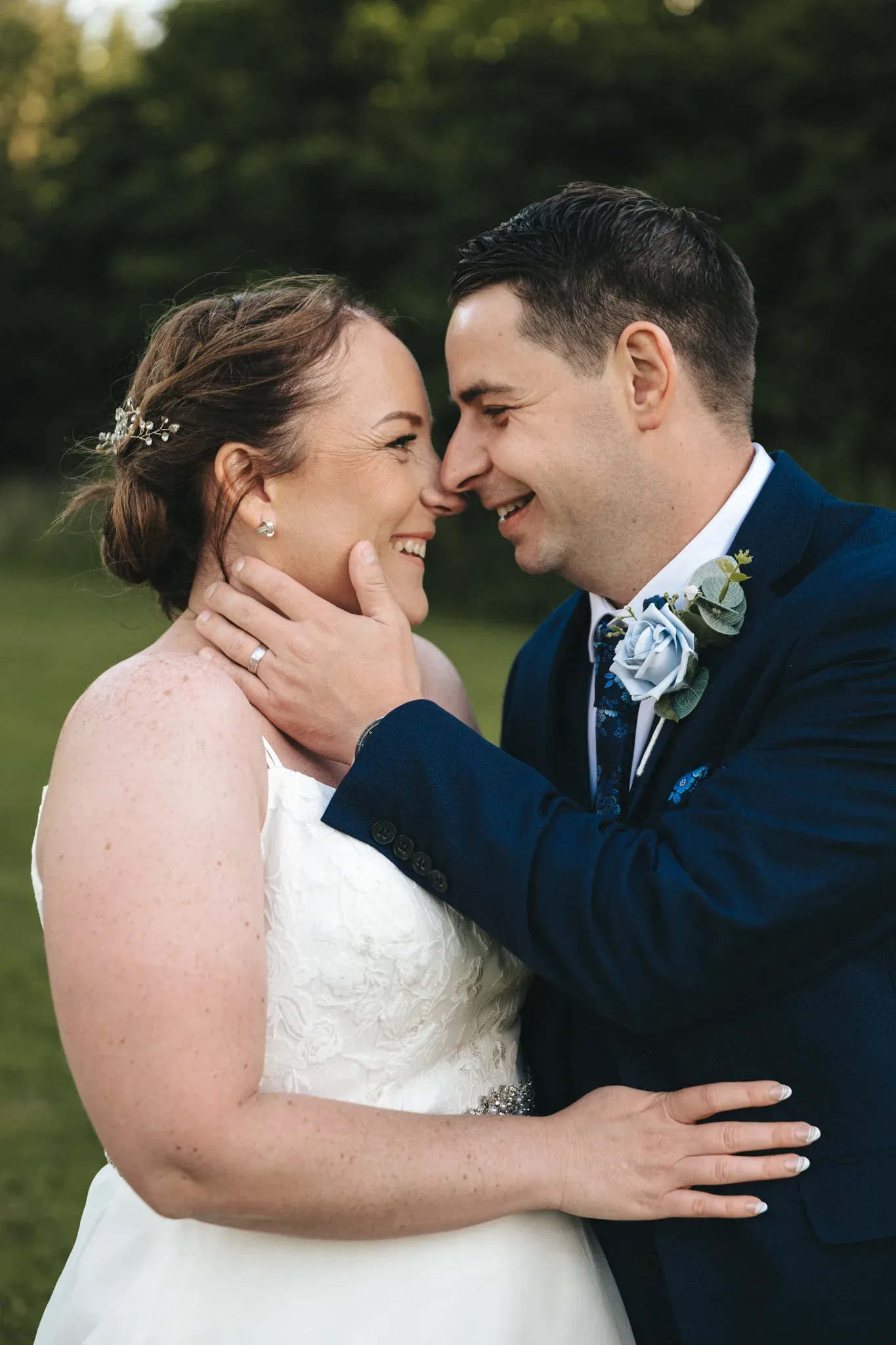 A bride and groom stand outside in a green, wooded area, close together and smiling. The bride is wearing a white wedding dress and has her hair styled with a decorative hairpiece. The groom is in a navy blue suit with a blue floral boutonniere, gently holding the bride's face with one hand. © Aimee Lince Photography